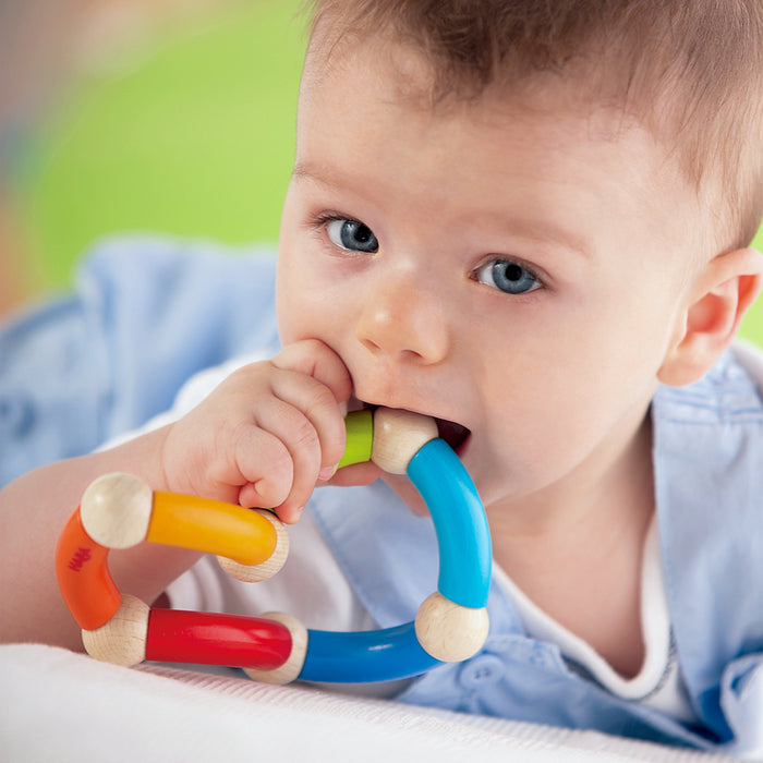 A blue-eyed baby with short hair in a light blue shirt enhances gross motor skills by chewing on HABA’s eco-friendly Color Snake Wooden Rattle & Fidget Toy featuring vibrant red, yellow, blue, and green sections against a softly blurred background.