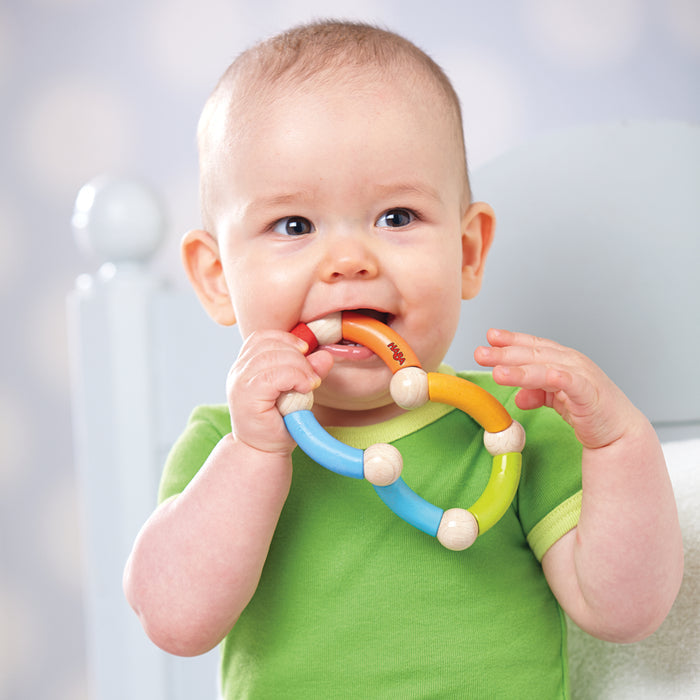 A baby in a green shirt chews on an eco-friendly HABA Color Snake Wooden Rattle & Fidget Toy while sitting on a light blue chair, set against a softly blurred background with circular light patterns, highlighting the focus on the babys developing gross motor skills.