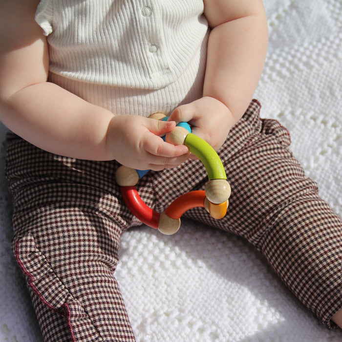 A baby sits on a textured white blanket, wearing a sleeveless top and checkered pants, while holding an eco-friendly Color Snake Wooden Rattle & Fidget Toy by HABA. This vibrant toy aids in gross motor skill development with its small, brightly colored connected segments.