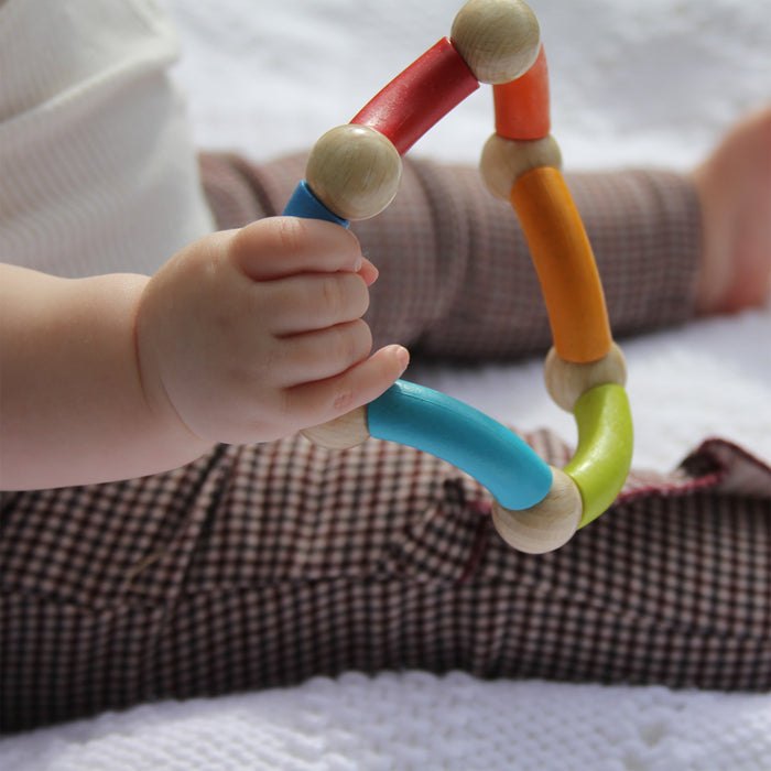 A baby in brown checkered pants sits on a white blanket, holding an eco-friendly HABA Color Snake Wooden Rattle & Fidget Toy. The vibrant segments—red, orange, blue, and green—are linked by round wooden beads to enhance gross motor skills.
