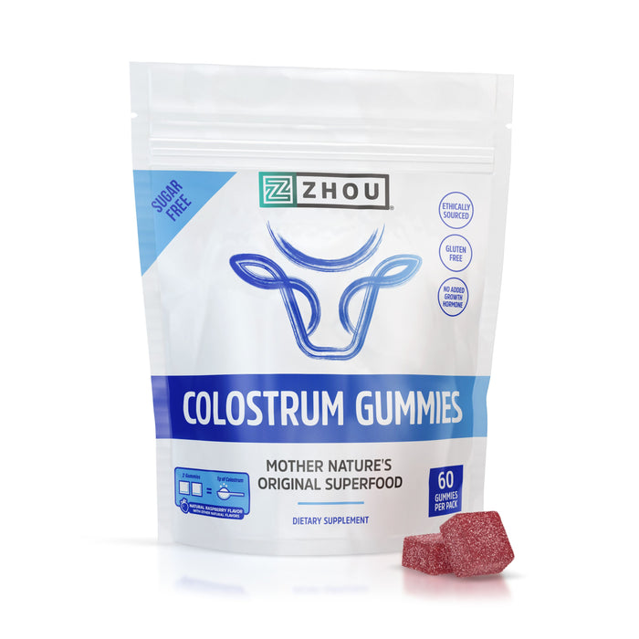 A Zhou Nutrition Colostrum Gummies Gummy package, featuring a white and blue design with a cow head graphic on a white background, is showcased with two red gummies beside it. The packaging highlights 60 superfood treats that are sugar-free and gluten-free.