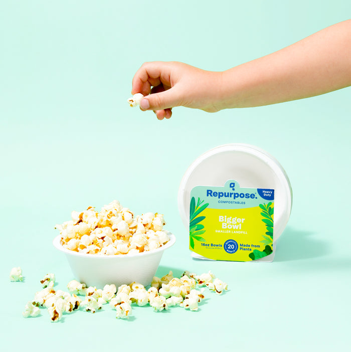 A hand reaches for popcorn in a Repurpose Compostable Bowl made from sugarcane scraps, surrounded by spilled kernels. A Repurpose biodegradable bowl lid is visible in the background against a light mint green backdrop.