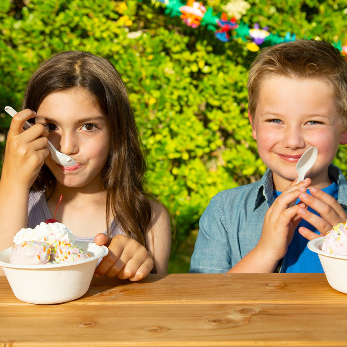 Two children sit enjoying ice cream in Repurpose Compostable Bowls, made from sugarcane scraps, at a wooden table. The girl on the left has a spoonful topped with whipped cream and sprinkles, while the boy on the right smiles as he holds his spoon. Green foliage frames their joyful moment.