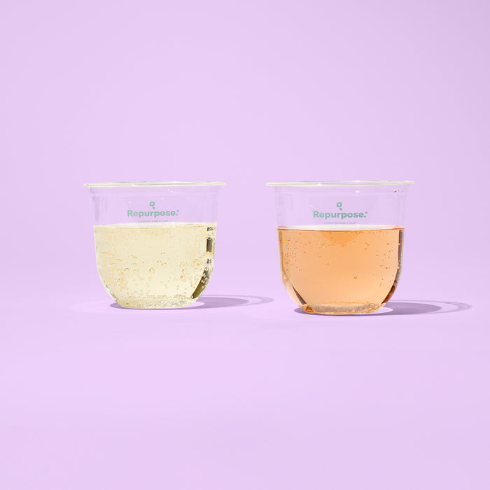 Two Repurpose Compostable Cold Cups, each featuring the brand logo, are shown on a pastel purple surface with one filled with a light yellow fizzy drink and the other with a light orange fizzy drink against a purple background.
