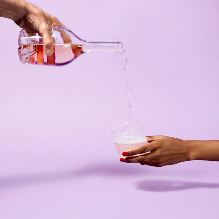 A person pours water from a glass carafe into a Repurpose Compostable Cold Cup, held by another person with red-painted nails, against a light purple background.