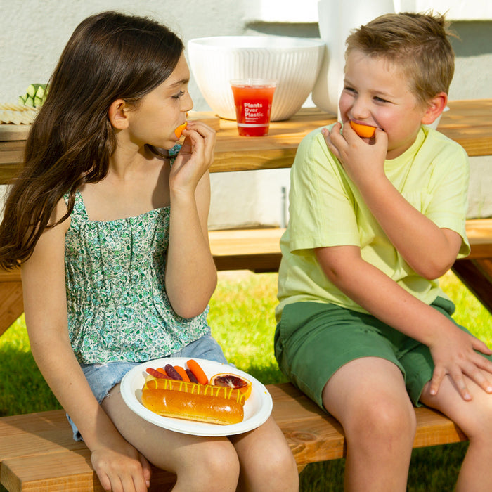 A girl and boy sit on a wooden bench outside, munching on carrot sticks. Shes in a green floral top and denim shorts, while hes in a light green shirt and shorts. On her lap is a Repurpose Compostable Dinner Paper Plate with a hot dog and carrots. A cup is visible in the background.