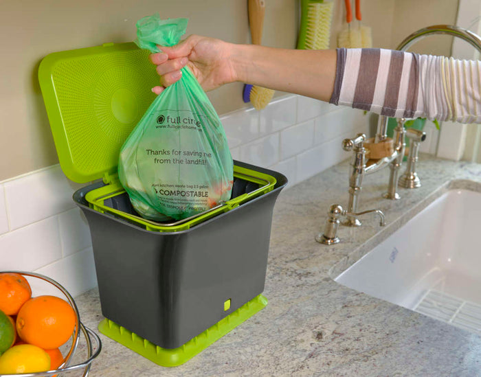Someone puts a Full Circle Compostable Food Scrap Bag in a black and green compost bin on the kitchen counter, underlined by its ASTM D6400 certification. Nearby, on the counter, a bowl of fruit is placed beside the sink with its silver faucet gleaming.