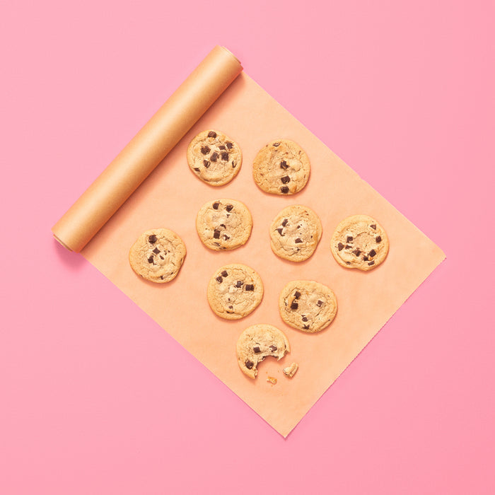 Ten chocolate chip cookies sit on Full Circles Compostable Parchment Paper Roll, with one cookie missing a tempting bite, set against a pink background.