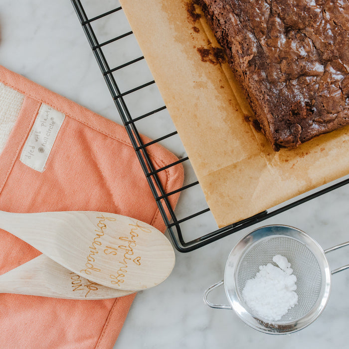 Brownies cool on Full Circles Compostable Parchment Paper Roll atop a black wire rack. Two wooden spoons, one with engraved text, sit on salmon fabric. A small sieve with powdered sugar lies nearby on marble.