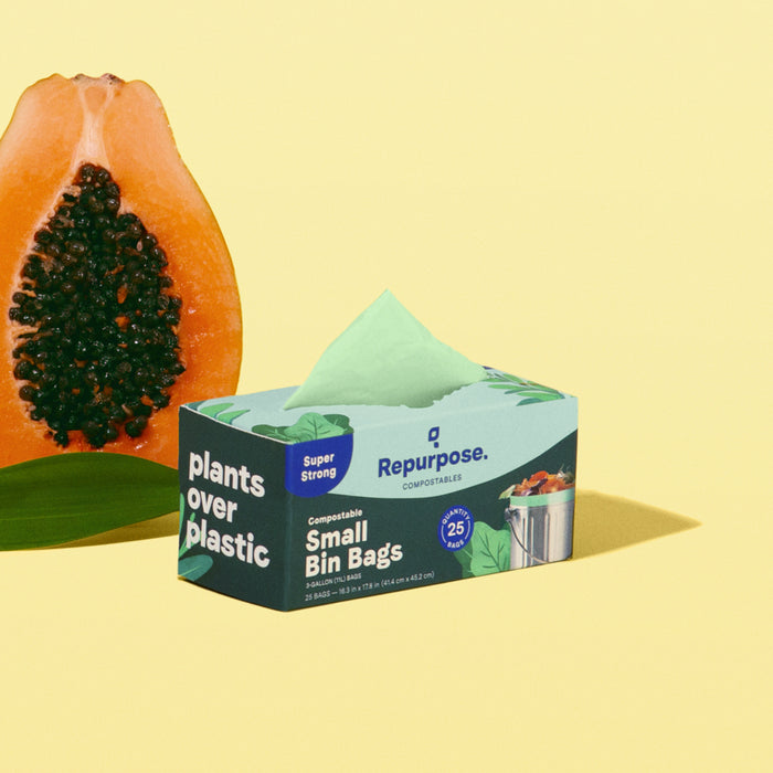 On a yellow background, a box of Repurpose Compostable Food Scrap Bags is shown beside a halved papaya. The box displays 25 non-toxic bags and features the Plants Over Plastic message, while the papayas orange flesh contrasts with its black seeds.