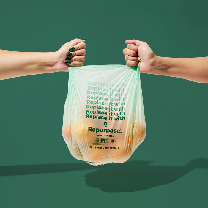 Two hands hold a translucent green Repurpose Compostable Small Bin Bag filled with oranges. The bag says “Replace it with Repurpose,” and is set against a solid green background.