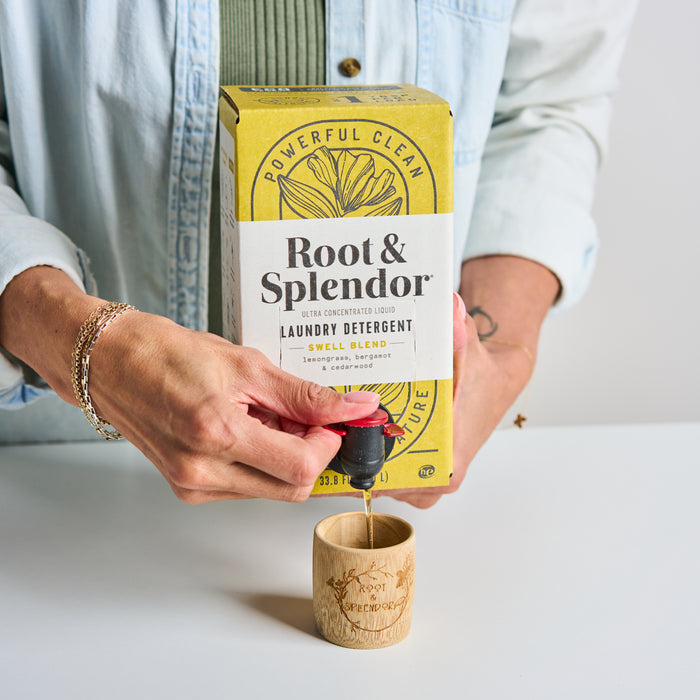 A person pours Root & Splendors Concentrated Laundry Detergent from a floral yellow box into a wooden cup. The box reads Ultra Concentrated Liquid Laundry Detergent, Swell Blend, Lavender, Bergamot, highlighting its eco-friendly formula and all-natural ingredients.
