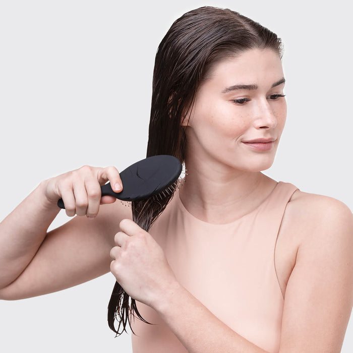 A woman with long brown hair uses the Kitsch Consciously Created Wet/Dry Brush, its flexible bristles smoothly gliding through her wet strands. She smiles slightly in a sleeveless beige top against a light gray background, embodying effortless hair care.