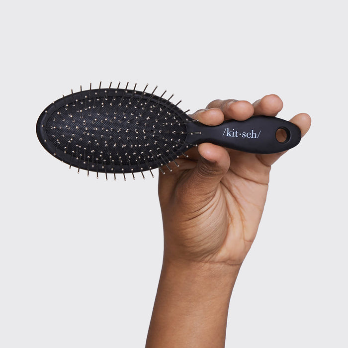 A hand holds Kitschs Consciously Created Wet/Dry Brush, a sleek black oval hairbrush with metallic flexible bristles, against a plain white background. The handle features the word kitsch and has a circular hole at the end.