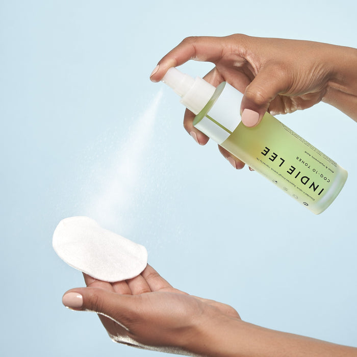A hand sprays Indie Lee CoQ10 Toner - Travel onto a white cotton pad against a light blue background, illustrating clean skincare in action.