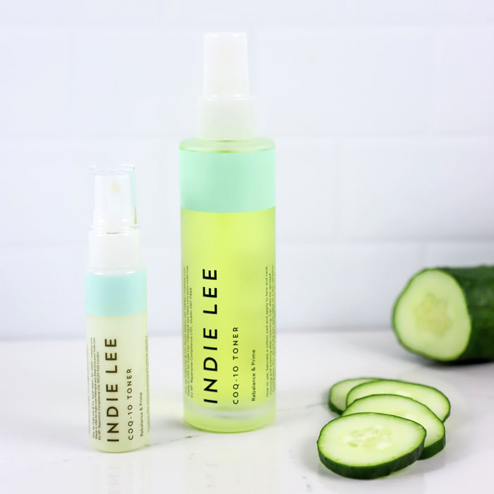 Two bottles of Indie Lee CoQ10 Toner - Travel, one small and one large, are displayed on a white surface beside cucumber slices and a partially visible cucumber with a white tiled background—ideal for your clean skincare routine.