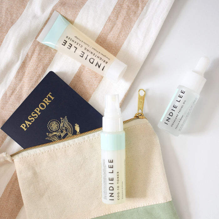 Flat lay of a striped pouch with a U.S. passport peeking out, next to three Indie Lee products—including the CoQ10 Toner - Travel—plus a gold zipper, all arranged on a white surface.