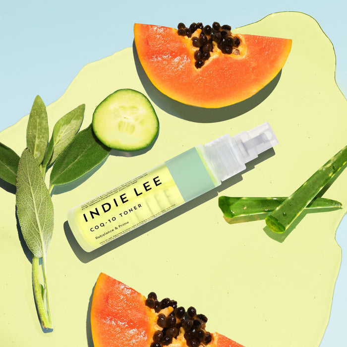 A bottle of Indie Lee CoQ10 Toner - Travel, a hydrating clean skincare favorite, is surrounded by papaya slices, cucumber, green leaves, and aloe vera on a light green background.