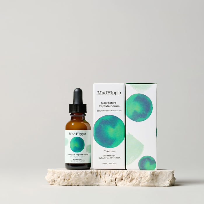 The Mad Hippie Corrective Peptide Serum bottle and its dropper are placed next to the packaging box, featuring abstract green and blue circular designs, all set elegantly on textured stone against a light gray background. Perfect for addressing sagging skin and wrinkles.