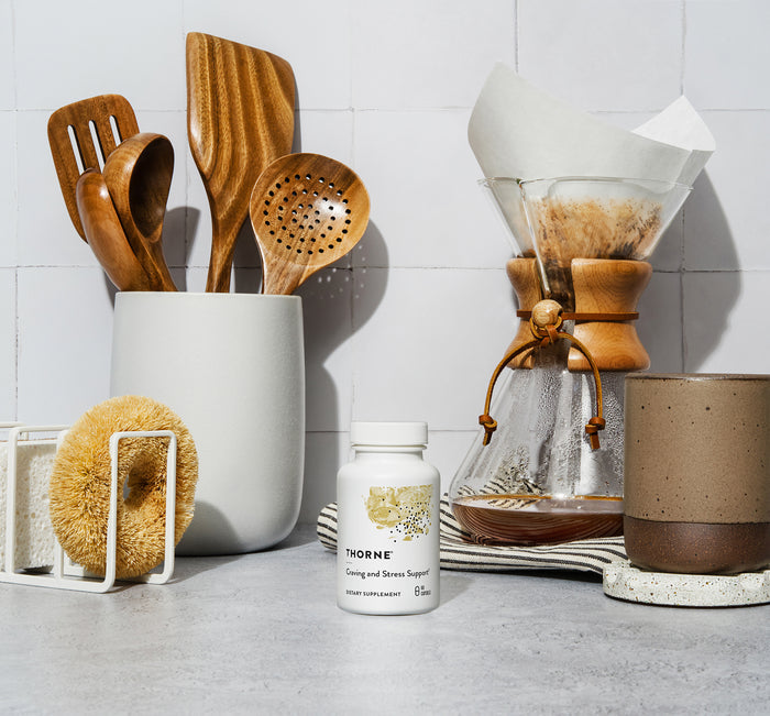 A kitchen counter displays a bottle of Thornes Craving and Stress Support supplements, wooden utensils in a holder, and a Chemex with filter. A brown mug and scrub brush add charm, while the tiled backsplash and textured coaster complete the serene setup.
