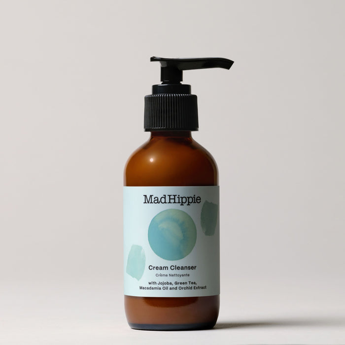 A minimalist brown pump bottle labeled Mad Hippie Cream Cleanser features paint swatches and text highlighting natural ingredients such as jojoba, green tea, macadamia oil, and orchid extract—all rich in antioxidants—set against a neutral background.