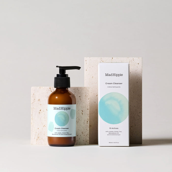 A Mad Hippie Cream Cleanser with a pump and minimalist design featuring a light blue circle sits beside its box on a soft stone-textured background, highlighting its antioxidant-rich, natural formula.