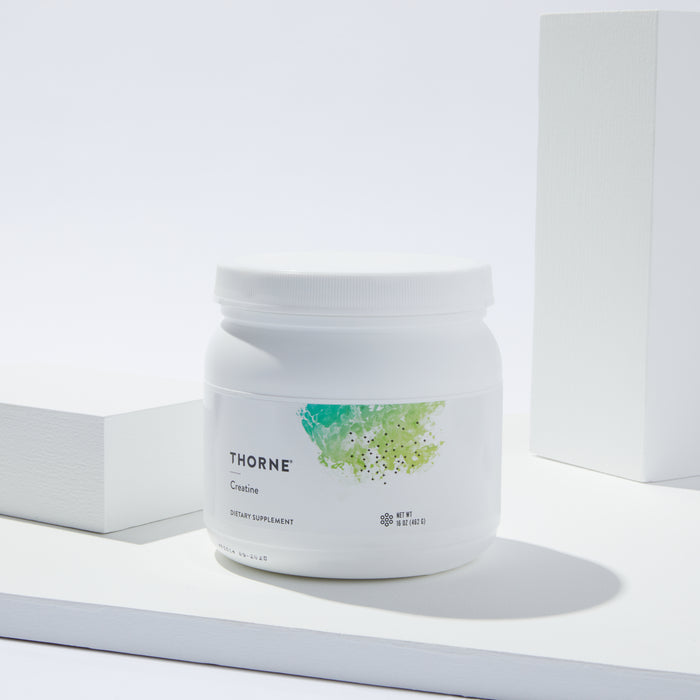 A white Thorne Creatine dietary supplement container rests on a sleek white surface with geometric blocks. Its label displays green abstract art, emphasizing its function in boosting cellular energy and supporting lean muscle mass.