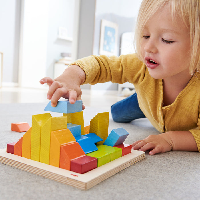 A child in a yellow shirt plays on a carpet, arranging colorful blocks from the HABA Creative Stones - 3D Wooden Arranging Blocks set, focusing intently as they add a blue block to the growing structure.