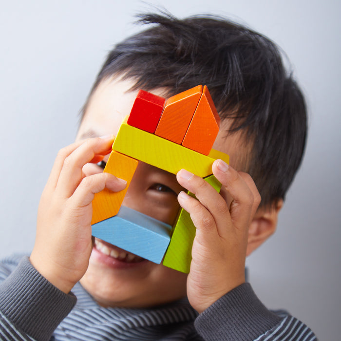 A playful child in a gray striped shirt smiles while holding HABAs Creative Stones - 3D Wooden Arranging Blocks, featuring vibrant colors like orange, red, yellow, green, and blue.