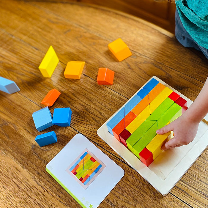 A child assembles a vibrant wooden puzzle on a wood surface, surrounded by colorful blocks. Nearby, a card displays the finished design of this HABA Creative Stones 3D Wooden Arranging Blocks masterpiece.