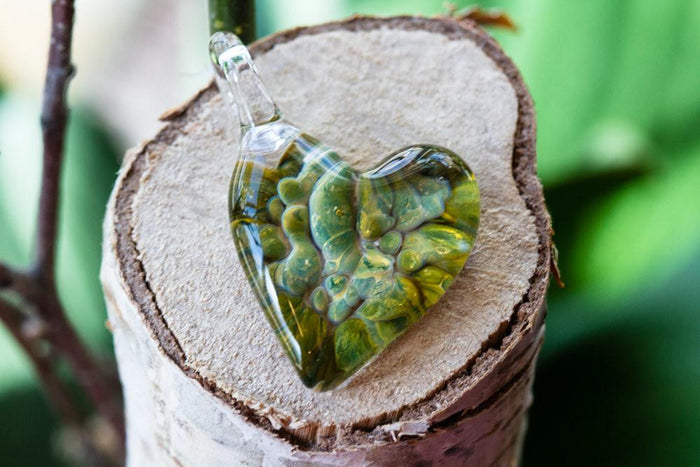 Green glass heart pendant with blue specks resting on a tree stump.