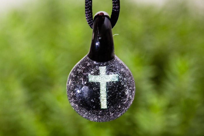 Close-up of round glass ash pendant with holographic cross, hung on a purple cord.