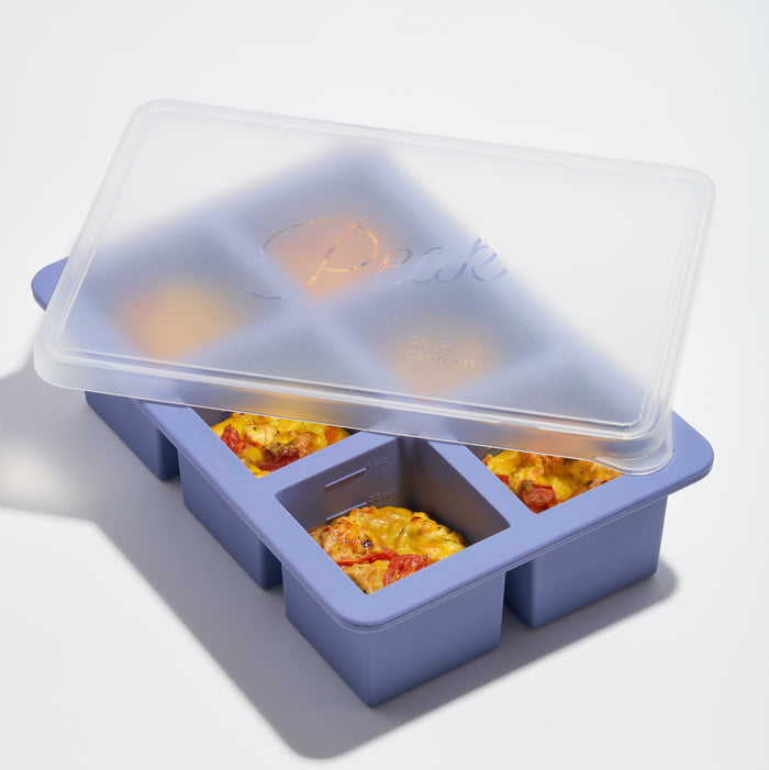 A partially open Cup Cubes Freezer Tray by W&P reveals three square portions of baked egg dishes with tomatoes and vegetables. The purple silicone tray, featuring a BPA-free lid, is highlighted on a white background, showcasing its practicality with soft lighting.
