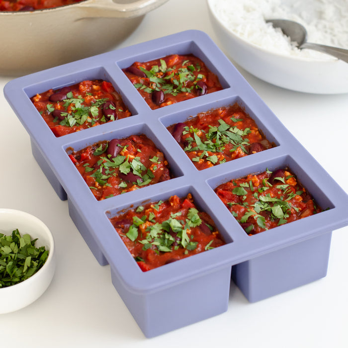 The W&P Cup Cubes Freezer Tray has six square silicone compartments filled with red chili topped with cilantro and comes with a BPA-free lid. In the background, theres a bowl of chopped cilantro and a plate of white rice to enhance your meal prep experience.
