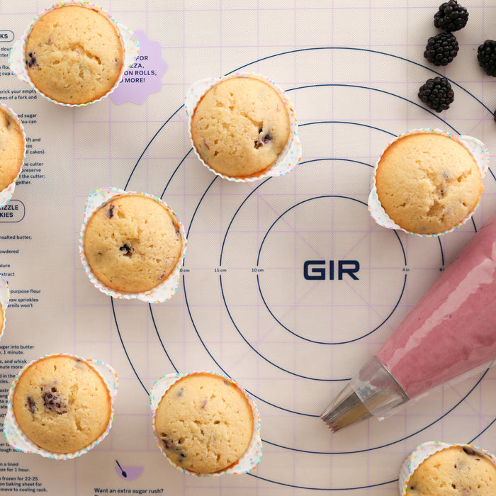 Top view of berry muffins in eco-friendly GIR Cupcake Liners on a pastry mat with circles. A piping bag of purple frosting sits beside them, and fresh blackberries are scattered in the upper right corner.