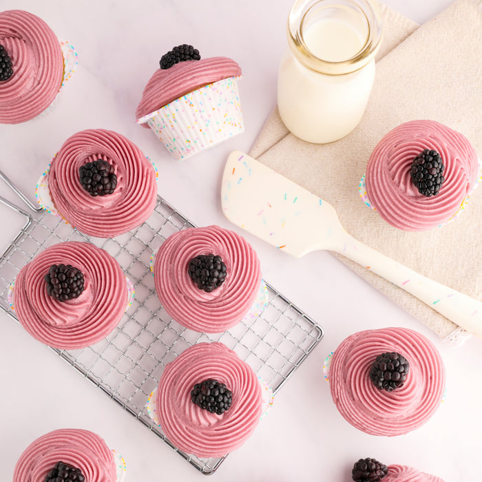 Pink-frosted cupcakes topped with blackberries rest snugly in eco-friendly GIR cupcake liners on a cooling rack and a marble surface. Nearby, a spatula sprinkled with colorful sprinkles and a small glass jar of milk on a beige cloth add charm to the delightful scene.