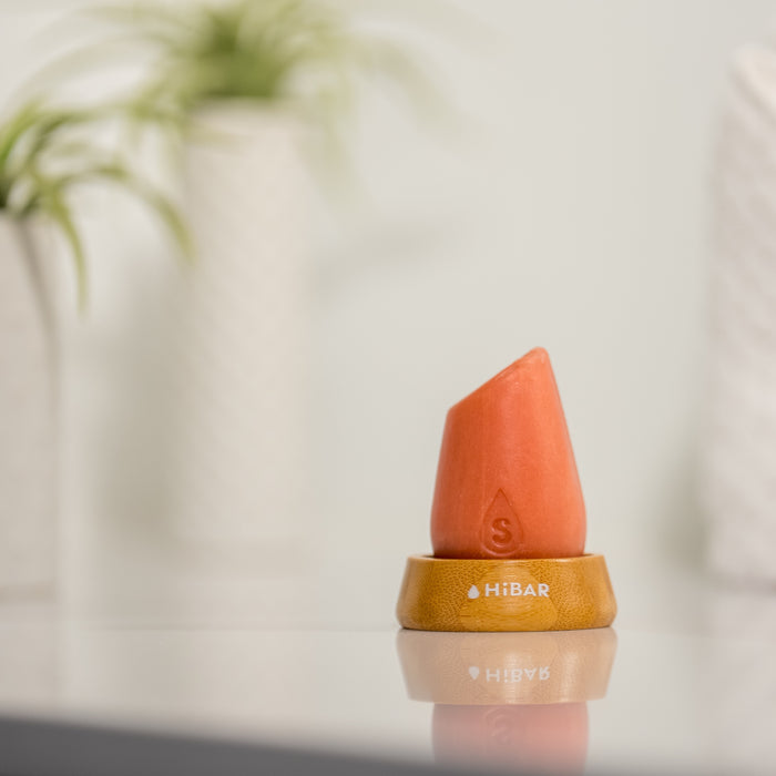 A coral-colored HiBAR Curl Shampoo Bar stands upright on a wooden base atop a reflective surface, with blurred green plants in white pots in the background. Rice protein enriches its nourishing formula, adding a touch of nature.