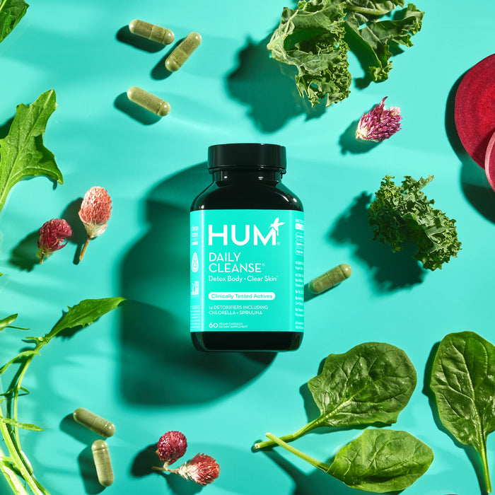 A bottle of HUM Nutrition Daily Cleanse Detox Supplement for clear skin, surrounded by leafy greens, herbs, and capsules on a teal background, promises powerful detoxifiers.