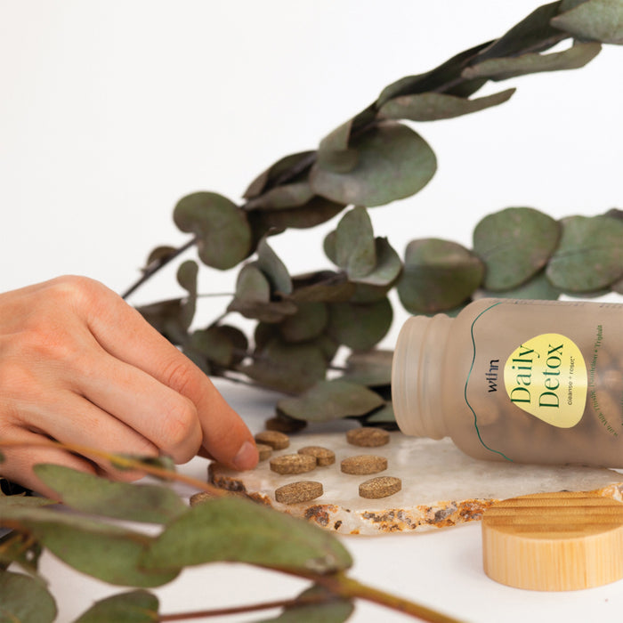 A hand reaches for supplements spilled from a Daily Detox - Cleanse + Reset bottle by WTHN on stone. Encircled by green eucalyptus leaves, it creates a natural, health-focused ambiance driven by an herbal blend of organic ingredients.