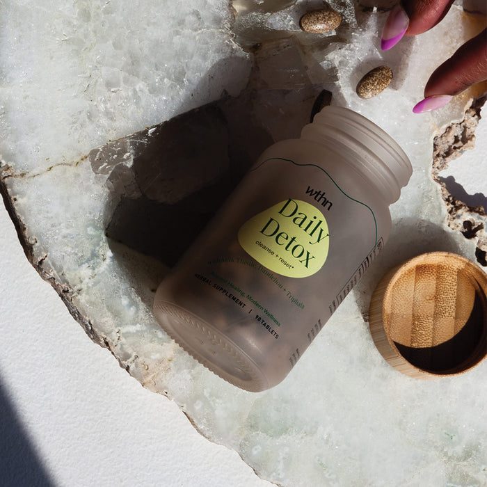 A hand with pink nails reaches for scattered tablets beside a Daily Detox - Cleanse + Reset glass jar by WTHN on stone. Made from organic ingredients, the herbal blend sits with its bamboo lid aside, as light casts shadows across the scene.