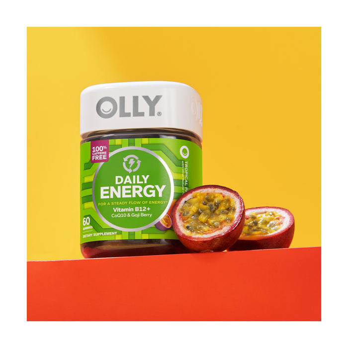 A container of OLLY Daily Energy vitamins with a green label is set against a vivid yellow and orange backdrop, with two halved passion fruits beside it. Rich in B12, these supplements provide the perfect daily energy boost.