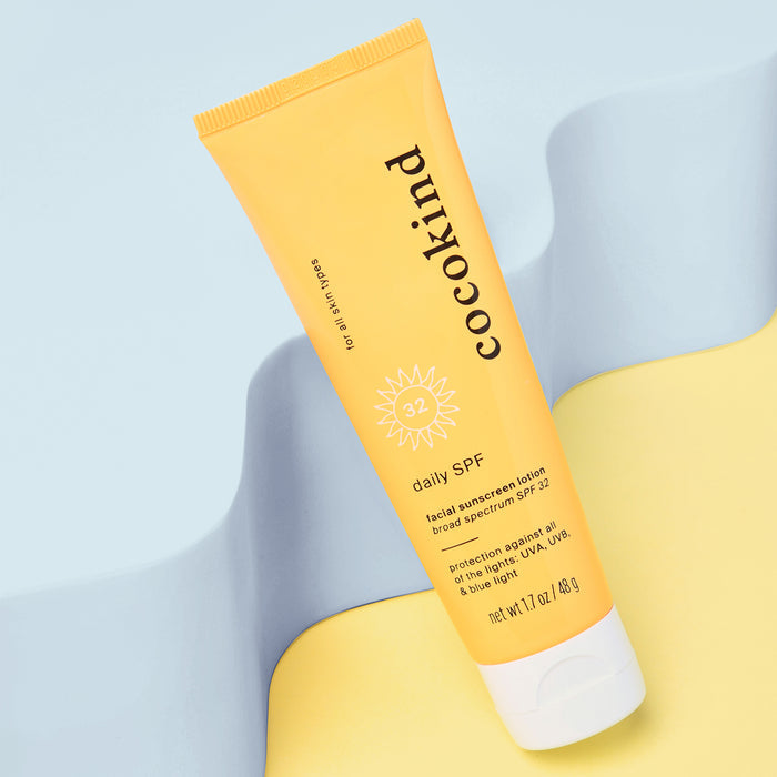 A yellow tube of Cocokinds Daily Facial Sunscreen Lotion with SPF 32 stands upright on a pastel yellow and blue background, featuring white text about its zinc oxide-enriched protection against UVA, UVB, and blue light.