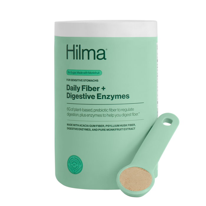 A pastel green cylindrical container labeled Daily Fiber + Digestive Enzymes by Hilma stands next to a scoop with beige powder. Ideal for sensitive stomachs, its label emphasizes plant-based prebiotic fiber, digestive enzymes, and no added sugar.