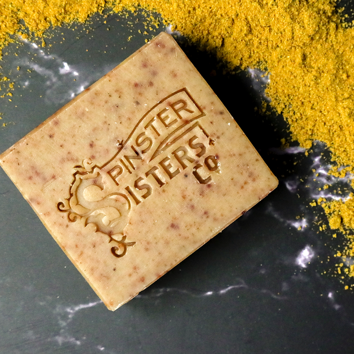 Spinster Sisters Co.s Face Cleanser Bar displays an intricate logo on a dark background. Nearby, bright turmeric powder adds contrast, while enriched with ingredients such as argan oil to provide indulgent skincare benefits.