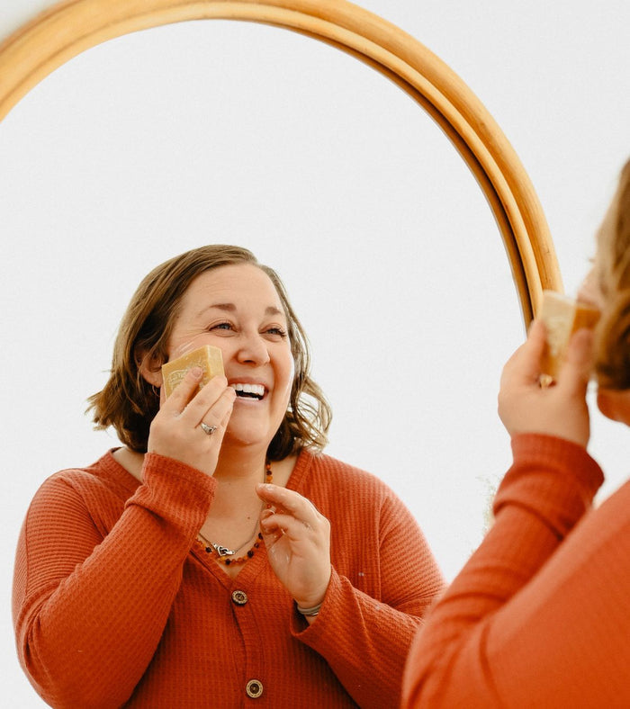A woman in a red shirt is smiling in front of a mirror, holding a Spinster Sisters Co. Face Cleanser Bar to her cheek. Her reflection is visible, and the setting appears bright and casual.