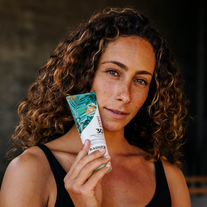 A woman with curly hair holds Badger Balms Daily Mineral Sunscreen - SPF 30, its tropical leaf design standing out. Clad in a black tank top and wearing a neutral expression, the blurred background emphasizes the focus on this essential moisturizer.