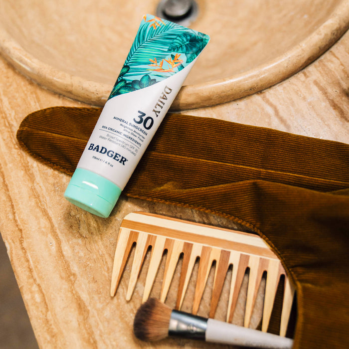 A tube of Badger Balms Daily Mineral Sunscreen - SPF 30 with a tropical design sits on a beige countertop alongside a wooden comb, makeup brush, and brown fabric item near the sink.