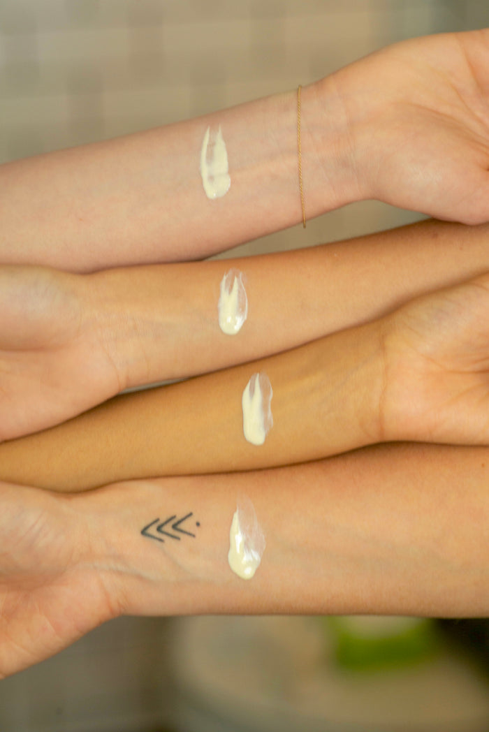 Three forearms of varying skin tones are aligned horizontally, each with a dab of Babo Botanicals Daily Sheer Fluid Mineral Sunscreen - SPF 50. One arm has an arrow tattoo, another wears a thin bracelet, while the background remains artfully blurred.