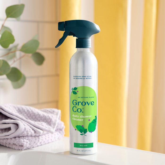 The Grove Co. Daily Shower Cleaner—Coming Soon, sits on a bathroom counter with purple towels. Its aluminum bottle has a green label and dark green nozzle, offering a plant-based formula. A yellow curtain and eucalyptus plant in the background create an inviting, shower-fresh atmosphere.