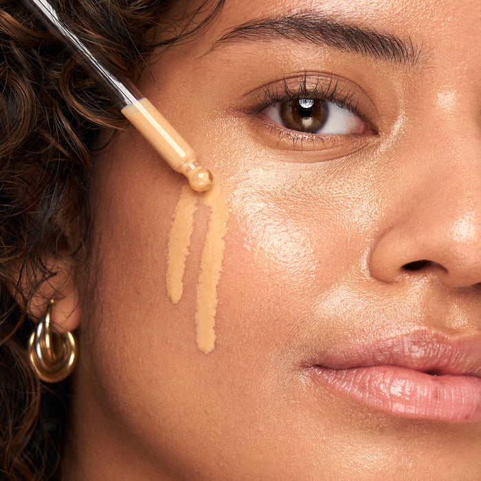 A close-up of a person with curly hair and gold hoop earrings shows them using Indie Lees Daily SPF Primer to apply foundation smoothly on their cheek, showcasing the products texture.